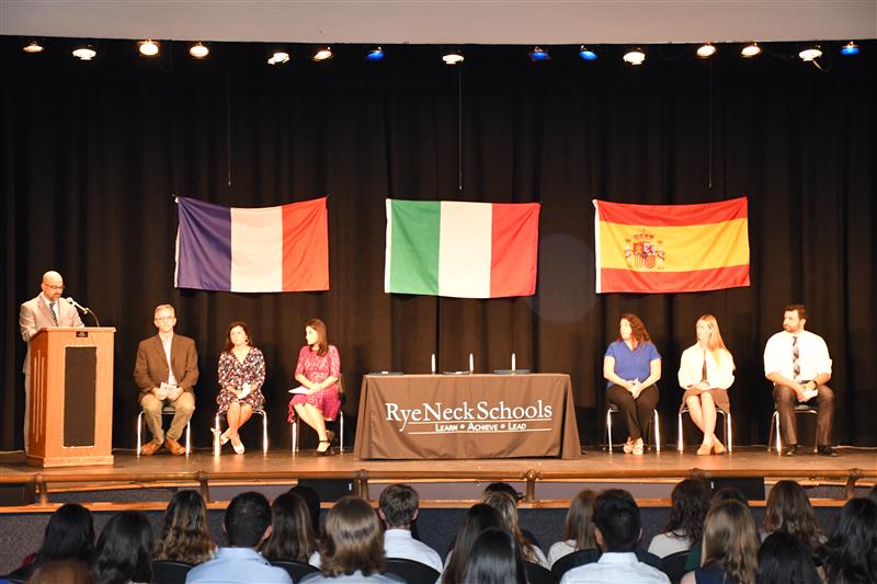 World Language teachers sit on stage while department chair speaks at a podium